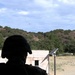 A silhouetted figure wearing a tactical/military helmet is observing from inside a building through an opening. In the background, there is an open training area with low structures, a solar panel on a pole, and a helicopter visible in the sky above a tree-covered hillside. The setting appears to be a military or law enforcement training facility.