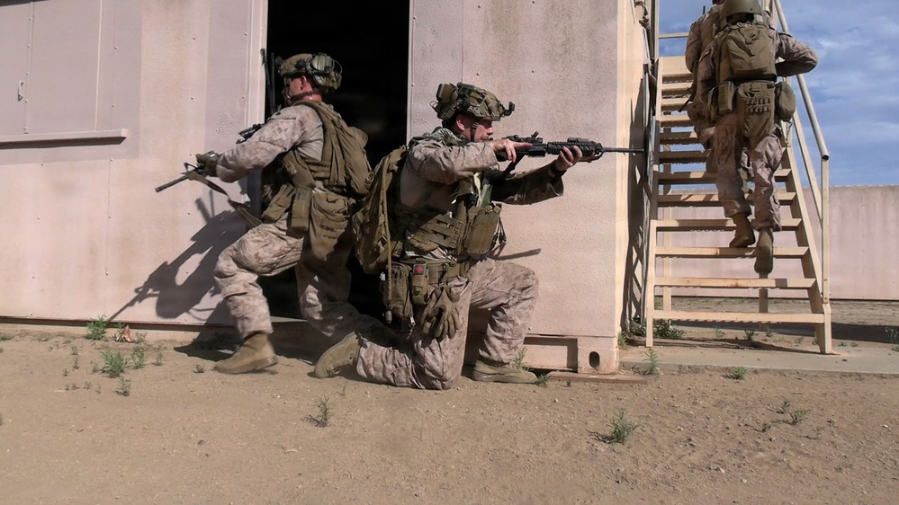 The image shows military personnel in desert camouflage uniforms conducting a tactical building clearance exercise at what appears to be a training facility. Three soldiers are visible: one kneeling and aiming a rifle outward providing cover, one entering a doorway, and one ascending an exterior staircase. The setting is a mock urban environment with sandy terrain, consistent with a military training range.