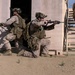 The image shows military personnel in desert camouflage uniforms conducting a tactical building clearance exercise at what appears to be a training facility. Three soldiers are visible: one kneeling and aiming a rifle outward providing cover, one entering a doorway, and one ascending an exterior staircase. The setting is a mock urban environment with sandy terrain, consistent with a military training range.