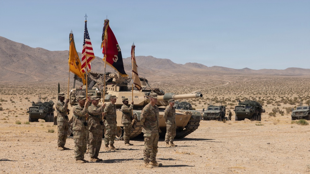 A U.S. Army military ceremony is taking place in an arid desert environment, likely at a training installation such as the National Training Center (NTC) at Fort Irwin, California. Soldiers in Army Combat Uniforms (ACU) are standing in formation holding multiple unit guidons and the American flag, with an M1 Abrams main battle tank and Stryker armored vehicles positioned behind them. The scene appears to be a formal change of command or unit activation ceremony conducted in the field.
