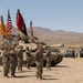 A U.S. Army military ceremony is taking place in an arid desert environment, likely at a training installation such as the National Training Center (NTC) at Fort Irwin, California. Soldiers in Army Combat Uniforms (ACU) are standing in formation holding multiple unit guidons and the American flag, with an M1 Abrams main battle tank and Stryker armored vehicles positioned behind them. The scene appears to be a formal change of command or unit activation ceremony conducted in the field.