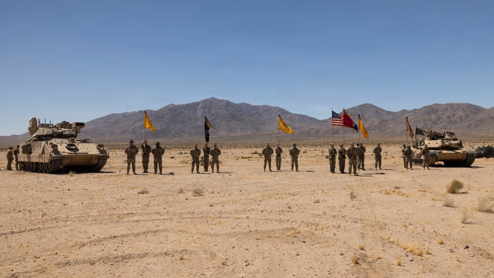 A group of U.S. Army soldiers are assembled in formation in an arid desert environment, flanked by two armored vehicles — an M2 Bradley Fighting Vehicle on the left and an M1 Abrams tank on the right. Multiple military guidons and the American flag are being displayed, suggesting a formal ceremony or unit photo. The setting appears to be a training range, likely the National Training Center (NTC) at Fort Irwin, California, based on the terrain and mountain backdrop.
