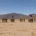 A group of U.S. Army soldiers are assembled in formation in an arid desert environment, flanked by two armored vehicles — an M2 Bradley Fighting Vehicle on the left and an M1 Abrams tank on the right. Multiple military guidons and the American flag are being displayed, suggesting a formal ceremony or unit photo. The setting appears to be a training range, likely the National Training Center (NTC) at Fort Irwin, California, based on the terrain and mountain backdrop.