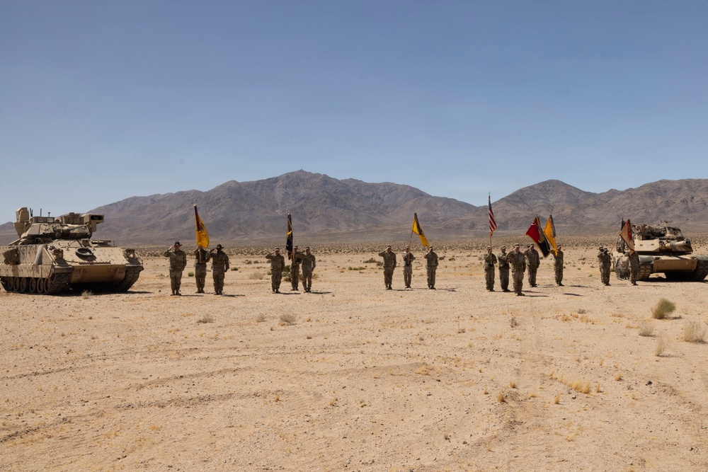 A military ceremony is taking place in an arid desert environment, with uniformed soldiers standing in formation and saluting while holding unit guidons and an American flag. Two armored vehicles flank the formation on either side, suggesting a formal change of command or awards ceremony. The backdrop features a clear blue sky and distant mountain range consistent with a Southwestern U.S. desert training installation.