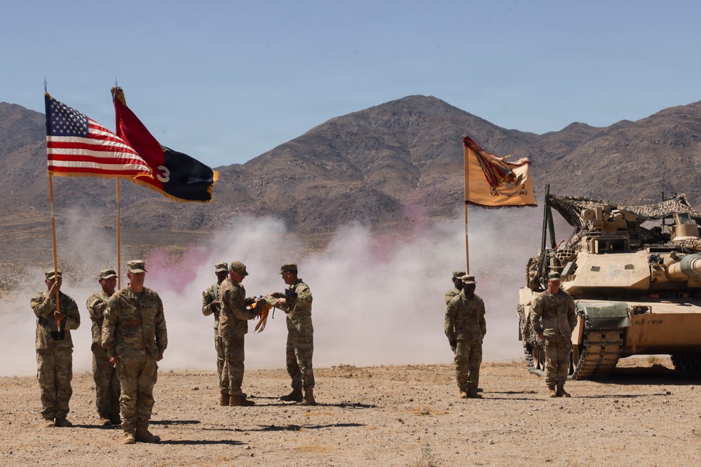 A U.S. Army military ceremony is taking place in a desert training environment, likely at the National Training Center (NTC) at Fort Irwin, California. Soldiers in Army Combat Uniforms (ACU) are participating in what appears to be a change of command or unit activation ceremony, with flags and a military vehicle present. Colored smoke grenades and dust create a dramatic backdrop against arid mountain terrain.