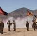 A U.S. Army military ceremony is taking place in a desert training environment, likely at the National Training Center (NTC) at Fort Irwin, California. Soldiers in Army Combat Uniforms (ACU) are participating in what appears to be a change of command or unit activation ceremony, with flags and a military vehicle present. Colored smoke grenades and dust create a dramatic backdrop against arid mountain terrain.
