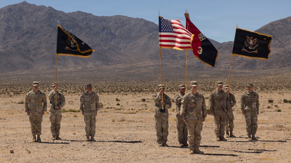 A group of U.S. Army soldiers are standing in formation in an arid desert landscape, holding multiple military flags including the American flag and unit colors. The ceremony appears to be a formal military event such as a change of command, activation, or unit ceremony. The setting features a stark desert terrain with mountain ranges visible in the background.