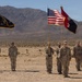 A group of U.S. Army soldiers are standing in formation in an arid desert landscape, holding multiple military flags including the American flag and unit colors. The ceremony appears to be a formal military event such as a change of command, activation, or unit ceremony. The setting features a stark desert terrain with mountain ranges visible in the background.
