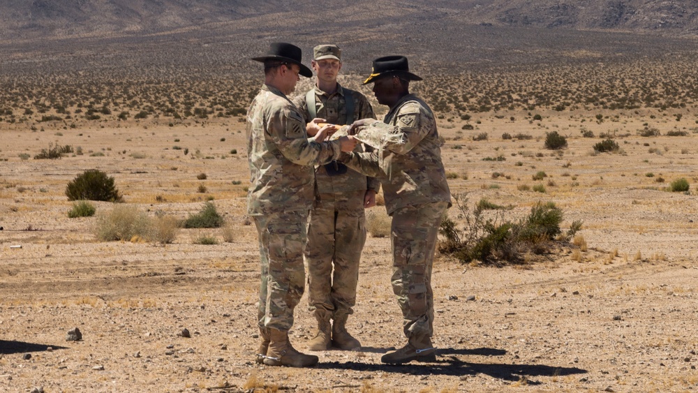 Three military personnel in U.S. Army MultiCam/OCP camouflage uniforms are standing in an arid desert environment. Two of the soldiers are wearing black cavalry-style Stetson hats, suggesting they are affiliated with a U.S. Army cavalry unit. The two foreground soldiers appear to be exchanging or examining an object together while a third soldier observes from behind.