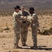 Three military personnel in U.S. Army MultiCam/OCP camouflage uniforms are standing in an arid desert environment. Two of the soldiers are wearing black cavalry-style Stetson hats, suggesting they are affiliated with a U.S. Army cavalry unit. The two foreground soldiers appear to be exchanging or examining an object together while a third soldier observes from behind.