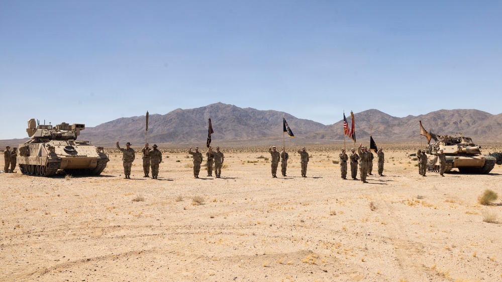 A military ceremony is taking place in an arid desert environment, with soldiers in formation saluting while holding multiple flags including the American flag. Two armored vehicles flank the formation on either side — a Bradley Fighting Vehicle on the left and an M1 Abrams tank on the right. The setting appears to be a training installation in a desert region, with mountain ranges visible in the background.
