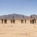 A military ceremony is taking place in an arid desert environment, with soldiers in formation saluting while holding multiple flags including the American flag. Two armored vehicles flank the formation on either side — a Bradley Fighting Vehicle on the left and an M1 Abrams tank on the right. The setting appears to be a training installation in a desert region, with mountain ranges visible in the background.