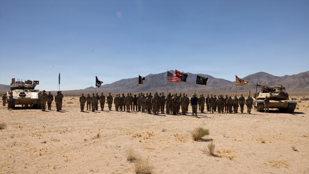 A large formation of U.S. Army soldiers stands at attention in an arid desert training environment, flanked by two armored vehicles. Multiple military unit flags and the American flag are displayed prominently behind the formation. The setting appears to be a formal ceremony or group photograph at a desert training installation.