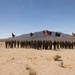A large formation of U.S. Army soldiers stands at attention in an arid desert training environment, flanked by two armored vehicles. Multiple military unit flags and the American flag are displayed prominently behind the formation. The setting appears to be a formal ceremony or group photograph at a desert training installation.