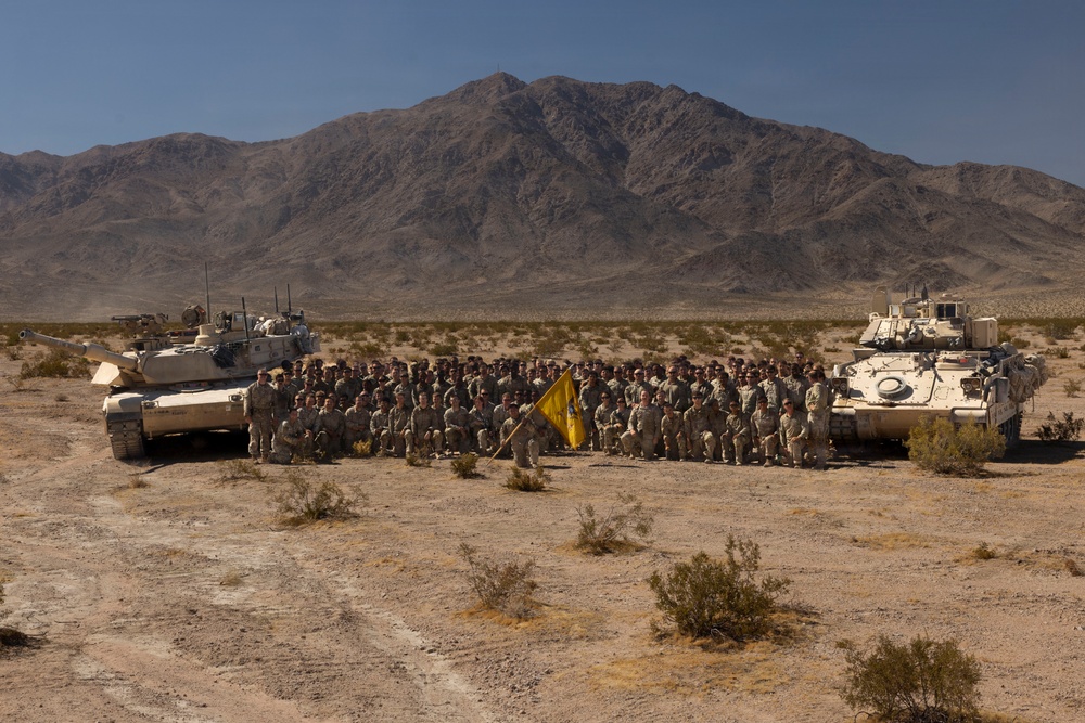 A large group of military personnel poses for a unit photograph in a desert environment flanked by two armored vehicles. The soldiers are arranged in rows between what appears to be an M1 Abrams main battle tank on the left and a Bradley Fighting Vehicle on the right. A yellow unit guidon flag is prominently displayed in the center of the formation.