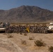 A large group of military personnel poses for a unit photograph in a desert environment flanked by two armored vehicles. The soldiers are arranged in rows between what appears to be an M1 Abrams main battle tank on the left and a Bradley Fighting Vehicle on the right. A yellow unit guidon flag is prominently displayed in the center of the formation.