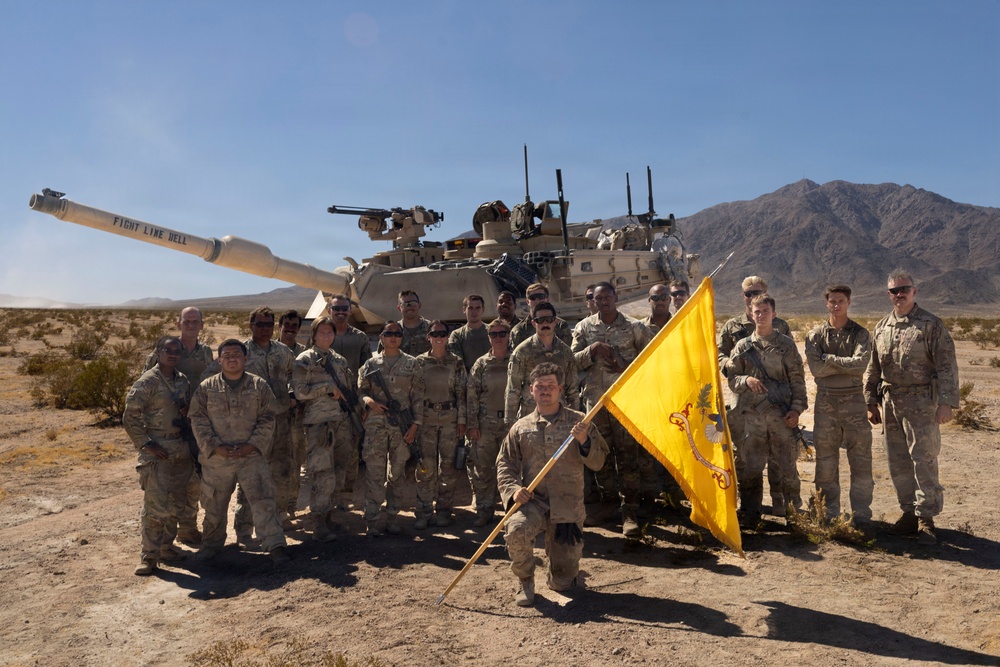 A group of approximately 20 U.S. Army soldiers in MultiCam/OCP camouflage uniforms pose for a unit photo in front of an M1 Abrams main battle tank in a desert environment. One soldier kneels in the front center holding a yellow unit guidon flag. The setting appears to be a desert training area, likely the National Training Center (NTC) at Fort Irwin, California, based on the terrain and mountain backdrop.
