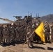 A group of approximately 20 U.S. Army soldiers in MultiCam/OCP camouflage uniforms pose for a unit photo in front of an M1 Abrams main battle tank in a desert environment. One soldier kneels in the front center holding a yellow unit guidon flag. The setting appears to be a desert training area, likely the National Training Center (NTC) at Fort Irwin, California, based on the terrain and mountain backdrop.