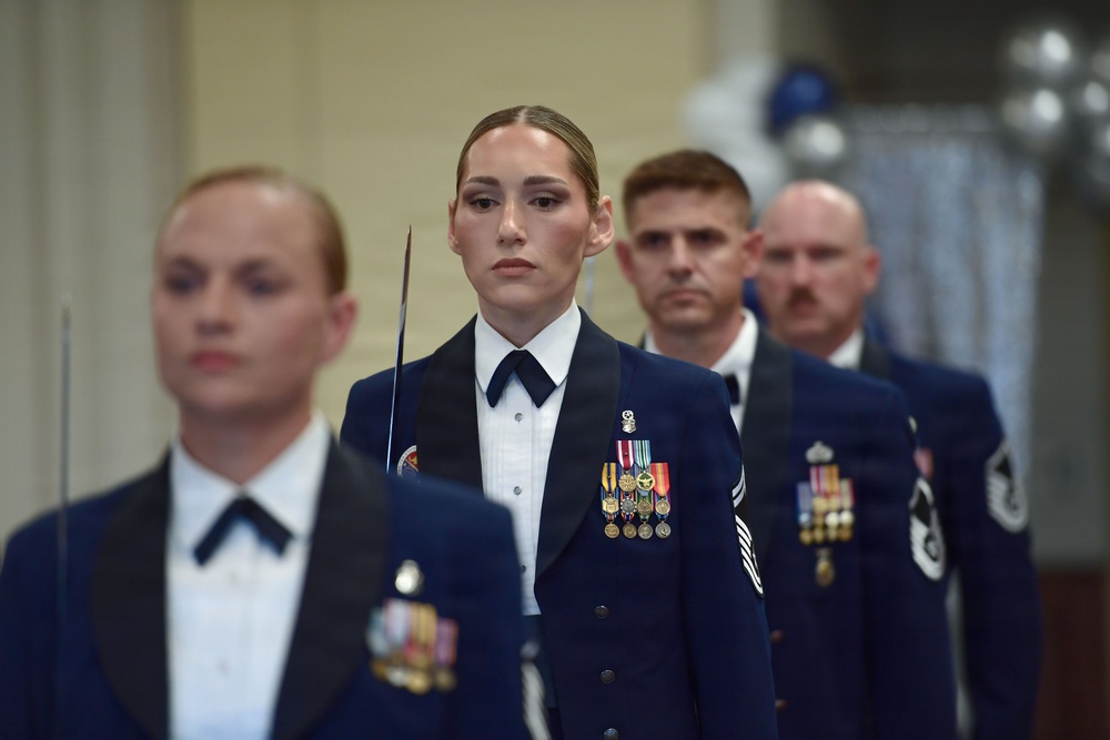 Four military personnel in formal dress uniforms are standing in a ceremonial formation, likely during an official military event or ceremony. They are wearing dark navy blue dress uniforms with white dress shirts, black bow ties, and multiple service medals and ribbons. The individuals maintain rigid, serious expressions consistent with a formal military ceremony.