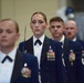Four military personnel in formal dress uniforms are standing in a ceremonial formation, likely during an official military event or ceremony. They are wearing dark navy blue dress uniforms with white dress shirts, black bow ties, and multiple service medals and ribbons. The individuals maintain rigid, serious expressions consistent with a formal military ceremony.