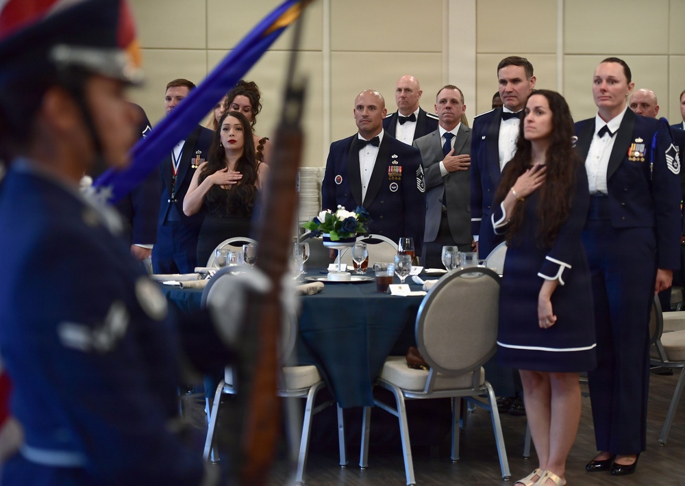 A formal military dining event, likely a military ball or formal dinner, is taking place in a banquet hall. Attendees in U.S. Air Force dress uniforms and formal civilian attire are standing at attention with hands over their hearts, indicating a pledge of allegiance or national anthem ceremony. A color guard member carrying a rifle and flag is visible in the foreground.