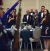A formal military dining event, likely a military ball or formal dinner, is taking place in a banquet hall. Attendees in U.S. Air Force dress uniforms and formal civilian attire are standing at attention with hands over their hearts, indicating a pledge of allegiance or national anthem ceremony. A color guard member carrying a rifle and flag is visible in the foreground.