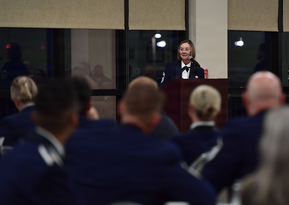 A woman in a dark military dress uniform stands at a wooden podium speaking into a microphone at what appears to be a formal military ceremony or event. She is addressing an audience of uniformed personnel seated with their backs to the camera. The setting appears to be an indoor venue with large windows visible in the background.