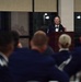 A woman in a dark military dress uniform stands at a wooden podium speaking into a microphone at what appears to be a formal military ceremony or event. She is addressing an audience of uniformed personnel seated with their backs to the camera. The setting appears to be an indoor venue with large windows visible in the background.