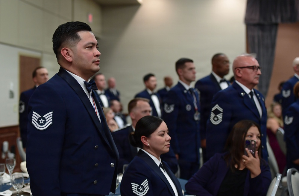A formal military ceremony or banquet featuring U.S. Air Force personnel in dress blue uniforms with bow ties. Multiple enlisted members are standing at attention in a banquet hall setting, with some civilians seated at tables. The rank insignia visible on uniforms indicates Technical Sergeant and Master Sergeant grades among the attendees.