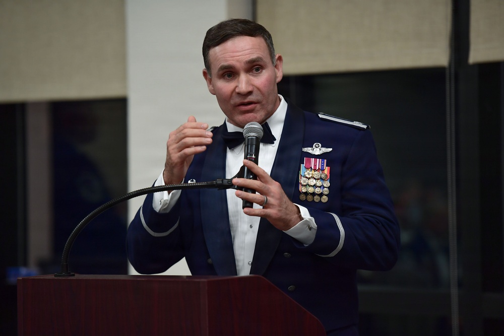 A U.S. Air Force officer in dress mess uniform is speaking at a podium while holding a microphone. He is wearing multiple service medals and ribbons, a master pilot badge, and silver shoulder boards indicating senior officer rank. The setting appears to be a formal military dining or awards ceremony.