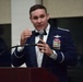 A U.S. Air Force officer in dress mess uniform is speaking at a podium while holding a microphone. He is wearing multiple service medals and ribbons, a master pilot badge, and silver shoulder boards indicating senior officer rank. The setting appears to be a formal military dining or awards ceremony.