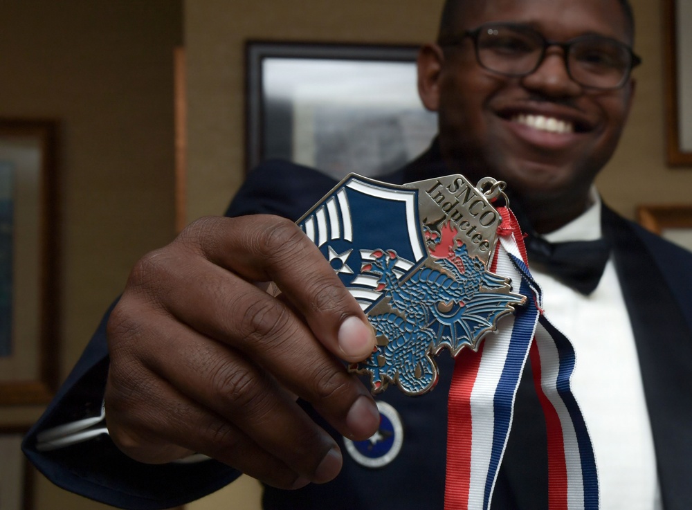 A smiling man in a dark blue uniform with a bow tie holds up a medallion toward the camera. The medallion reads 'SNCO Inductee' and features an eagle design with a blue and red enamel shield, attached to a red, white, and blue ribbon. The image appears to document a military induction ceremony, likely for the U.S. Air Force Senior Non-Commissioned Officer (SNCO) corps.
