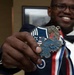 A smiling man in a dark blue uniform with a bow tie holds up a medallion toward the camera. The medallion reads 'SNCO Inductee' and features an eagle design with a blue and red enamel shield, attached to a red, white, and blue ribbon. The image appears to document a military induction ceremony, likely for the U.S. Air Force Senior Non-Commissioned Officer (SNCO) corps.