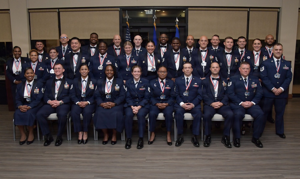 A formal group portrait of approximately 35 United States Air Force personnel in dress blue uniforms, all wearing red-white-and-blue ribbon medals around their necks. The group is arranged in three rows — seated, standing middle, and standing back — in what appears to be an indoor ceremonial venue. Two flags (American and Air Force) are visible in the background, suggesting this is an official recognition or graduation ceremony.