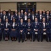 A formal group portrait of approximately 35 United States Air Force personnel in dress blue uniforms, all wearing red-white-and-blue ribbon medals around their necks. The group is arranged in three rows — seated, standing middle, and standing back — in what appears to be an indoor ceremonial venue. Two flags (American and Air Force) are visible in the background, suggesting this is an official recognition or graduation ceremony.