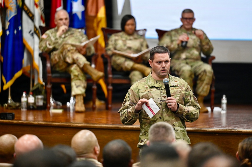 A U.S. Air Force officer named Taylor, identifiable by his name tape and 'U.S. Air Force' chest tape, stands before an audience holding a microphone and index cards, apparently delivering a presentation or asking a question. Behind him on a raised wooden stage, three military personnel in OCP (Operational Camouflage Pattern) uniforms are seated in chairs, suggesting a panel or senior leadership forum. Multiple military flags are displayed on the stage backdrop.