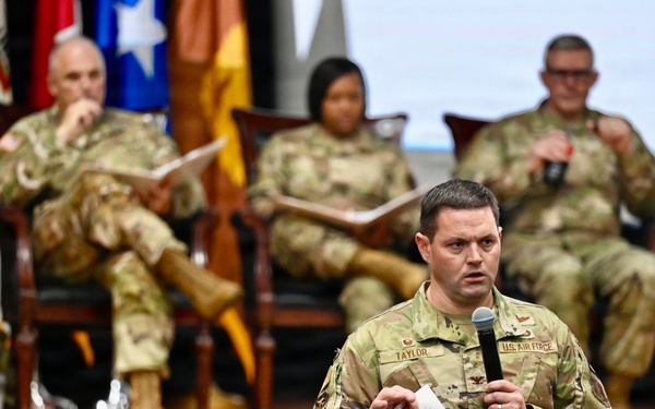 A U.S. Air Force officer named Taylor, identifiable by his name tape and 'U.S. Air Force' chest tape, stands before an audience holding a microphone and index cards, apparently delivering a presentation or asking a question. Behind him on a raised wooden stage, three military personnel in OCP (Operational Camouflage Pattern) uniforms are seated in chairs, suggesting a panel or senior leadership forum. Multiple military flags are displayed on the stage backdrop.