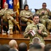 A U.S. Air Force officer named Taylor, identifiable by his name tape and 'U.S. Air Force' chest tape, stands before an audience holding a microphone and index cards, apparently delivering a presentation or asking a question. Behind him on a raised wooden stage, three military personnel in OCP (Operational Camouflage Pattern) uniforms are seated in chairs, suggesting a panel or senior leadership forum. Multiple military flags are displayed on the stage backdrop.