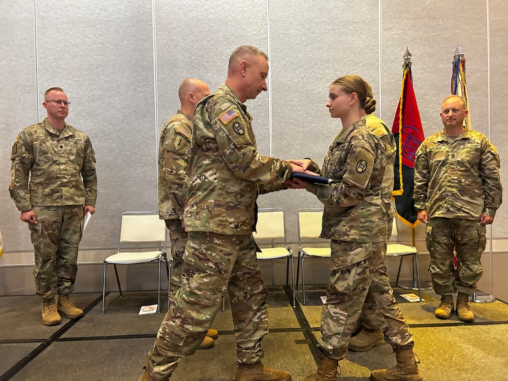 A U.S. Army military ceremony is taking place in an indoor venue where a senior officer is presenting a folder or award to a female soldier. Four soldiers in OCP (Operational Camouflage Pattern) uniforms are visible, with two standing at attention in the background. Military unit flags and guidons are displayed in the background.