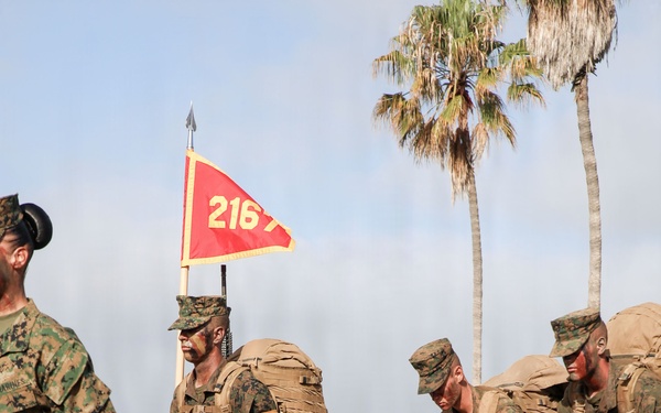 A group of U.S. Marines in MARPAT camouflage uniforms conducting a formation march or hike on a paved road. The lead Marine carries a red guidon pennant marked '216', and several recruits or junior Marines carry large rucksacks and M16/M4-style rifles. The setting appears to be a military base, likely on the West Coast based on the palm trees and coastal haze visible in the background.