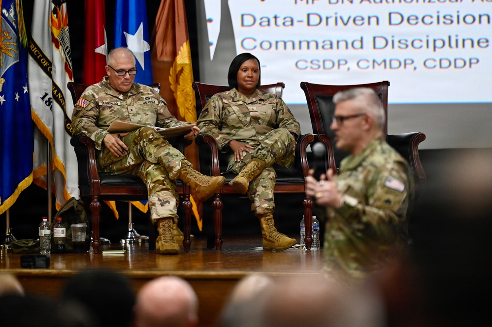 Three U.S. Army soldiers are on a stage during what appears to be a military briefing or conference. Two soldiers are seated in formal chairs on stage while a third soldier in the foreground appears to be speaking or presenting. A presentation screen in the background displays text related to 'Data-Driven Decision' and 'Command Discipline' with acronyms CSDP, CMDP, and CDDP.