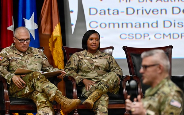 Three U.S. Army soldiers are on a stage during what appears to be a military briefing or conference. Two soldiers are seated in formal chairs on stage while a third soldier in the foreground appears to be speaking or presenting. A presentation screen in the background displays text related to 'Data-Driven Decision' and 'Command Discipline' with acronyms CSDP, CMDP, and CDDP.