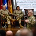 Three U.S. Army soldiers are on a stage during what appears to be a military briefing or conference. Two soldiers are seated in formal chairs on stage while a third soldier in the foreground appears to be speaking or presenting. A presentation screen in the background displays text related to 'Data-Driven Decision' and 'Command Discipline' with acronyms CSDP, CMDP, and CDDP.