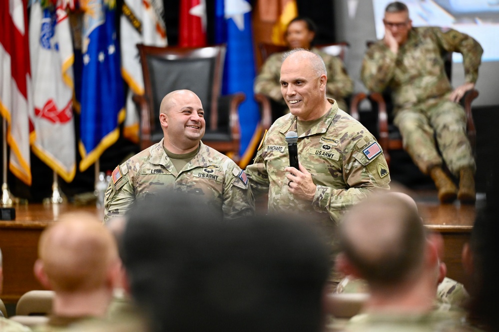Two U.S. Army soldiers stand at the front of a military auditorium or assembly hall, with one holding a microphone and speaking to an audience of uniformed personnel. Both soldiers are smiling, suggesting a positive or celebratory event such as an award ceremony, promotion, or unit gathering. Multiple military flags and additional soldiers are visible in the background.