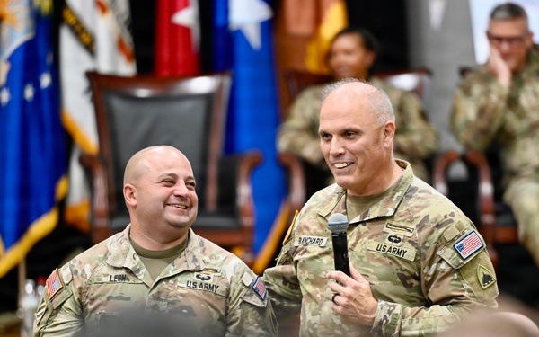 Two U.S. Army soldiers stand at the front of a military auditorium or assembly hall, with one holding a microphone and speaking to an audience of uniformed personnel. Both soldiers are smiling, suggesting a positive or celebratory event such as an award ceremony, promotion, or unit gathering. Multiple military flags and additional soldiers are visible in the background.