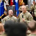 Two U.S. Army soldiers stand at the front of a military auditorium or assembly hall, with one holding a microphone and speaking to an audience of uniformed personnel. Both soldiers are smiling, suggesting a positive or celebratory event such as an award ceremony, promotion, or unit gathering. Multiple military flags and additional soldiers are visible in the background.