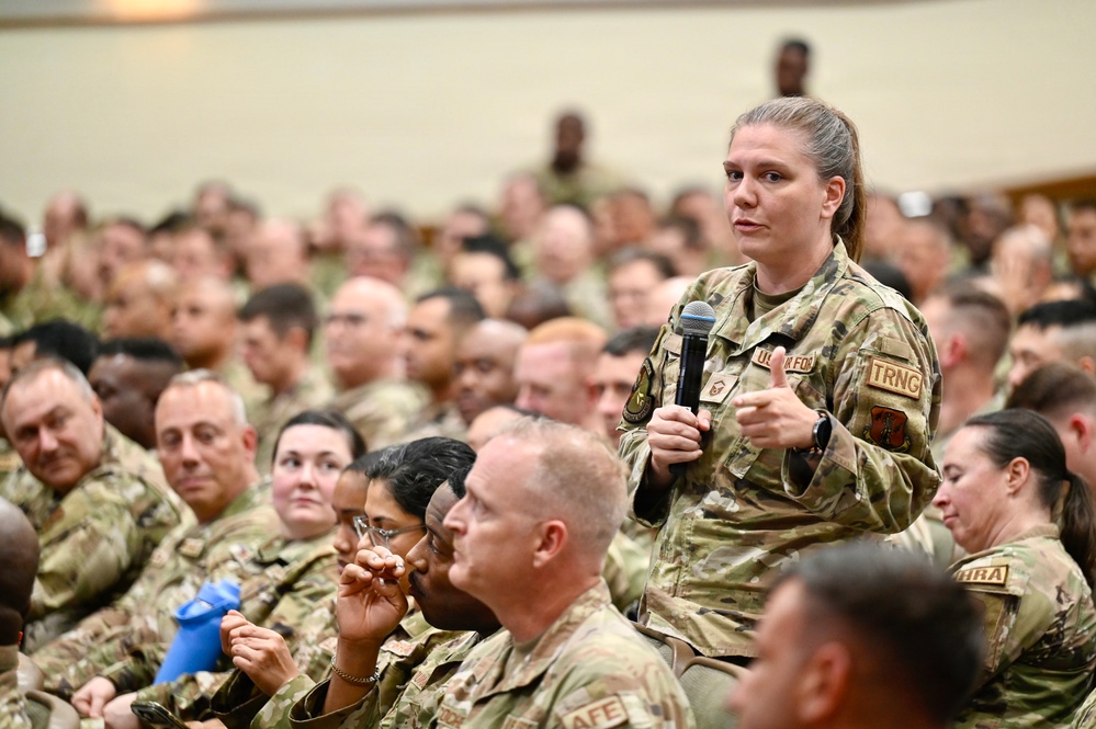 A large gathering of U.S. Air Force personnel in a auditorium or assembly hall setting. A female service member is standing and speaking into a microphone, likely during a question-and-answer session or all-hands meeting. The audience consists of hundreds of uniformed military personnel seated in rows.