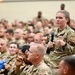 A large gathering of U.S. Air Force personnel in a auditorium or assembly hall setting. A female service member is standing and speaking into a microphone, likely during a question-and-answer session or all-hands meeting. The audience consists of hundreds of uniformed military personnel seated in rows.