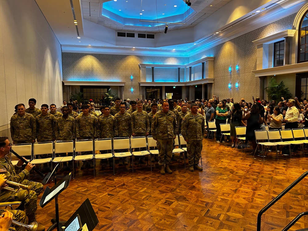 A military ceremony is taking place in a formal ballroom or event hall, where a line of soldiers in camouflage uniforms stands at attention in front of rows of chairs. A live military band is visible in the foreground on the left, and a large civilian audience is gathered on the right side observing the event. The venue features an ornate ceiling with blue accent lighting, suggesting a formal official occasion such as a graduation, promotion, or enlistment ceremony.