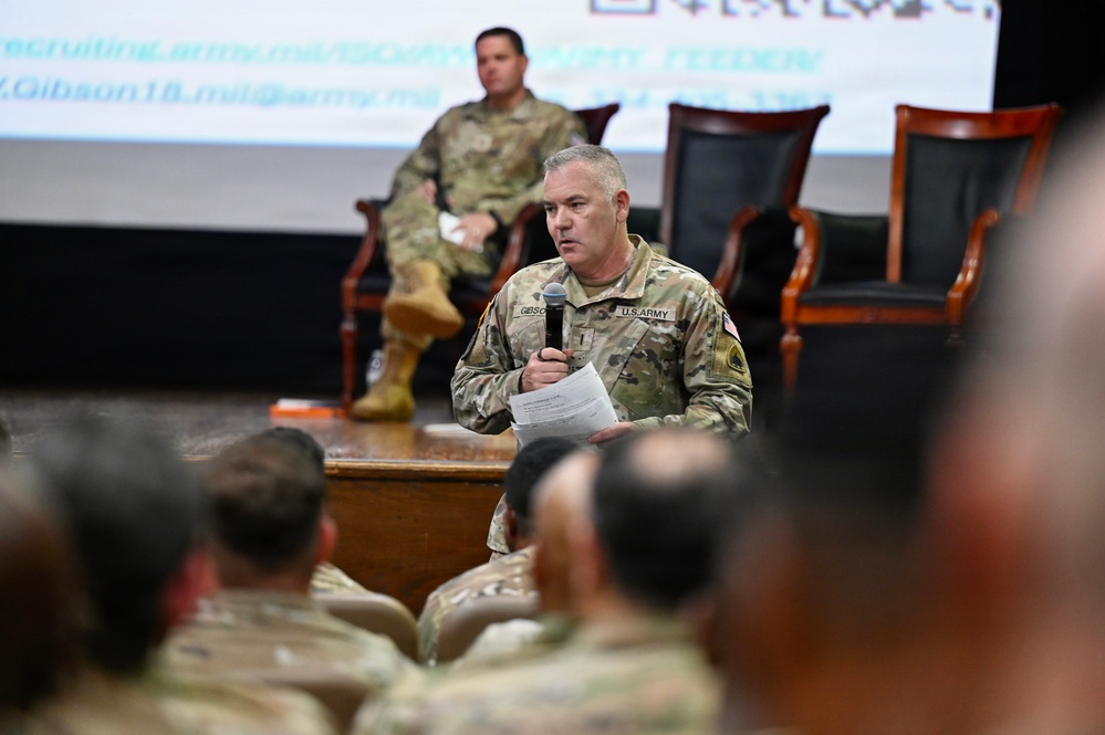 A U.S. Army soldier in OCP (Operational Camouflage Pattern) uniform is standing at the front of an auditorium-style room, speaking into a microphone while holding papers. A second soldier is seated on a chair on a raised stage in the background. The event appears to be a military briefing or town hall, with an audience of uniformed personnel visible in the foreground.